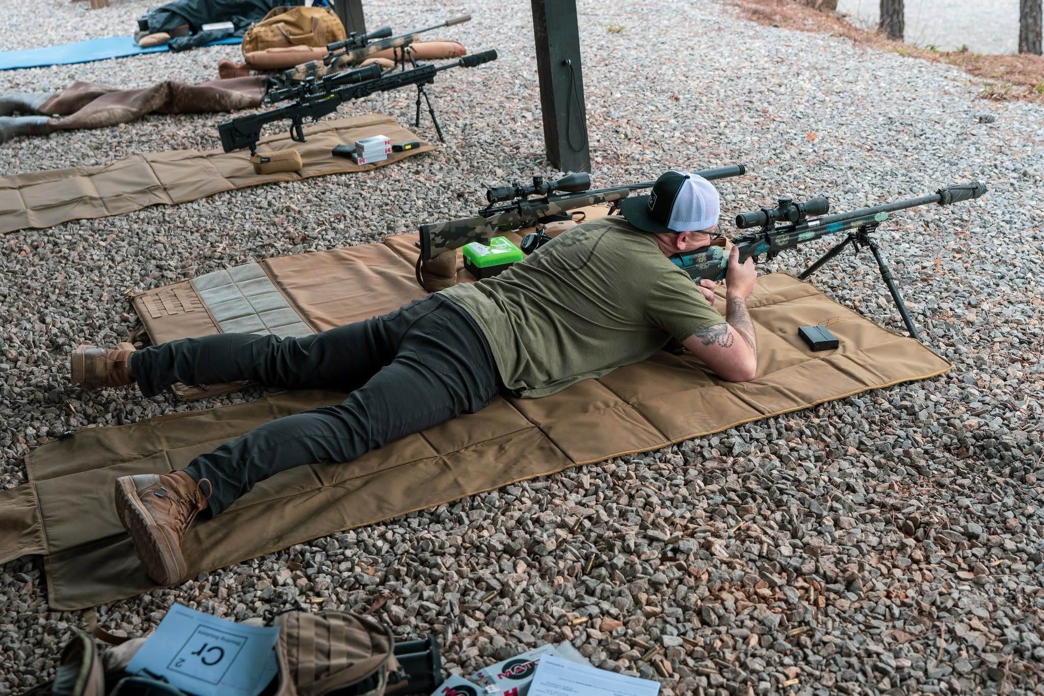 Shooter in prone position aiming a scoped rifle from a shooting mat on gravel during training